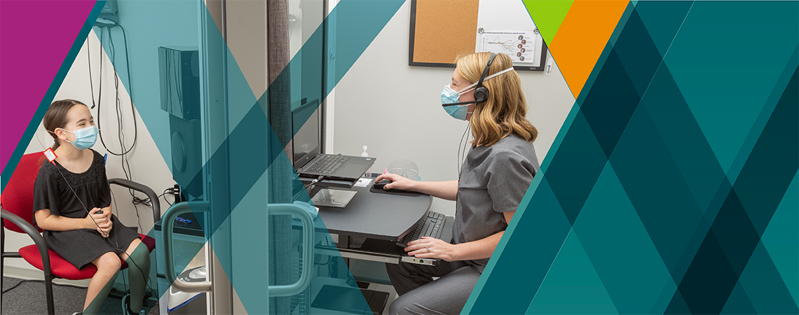 An audiologist sitting at a laptop tests a young girl's hearing. Both wear surgical masks.